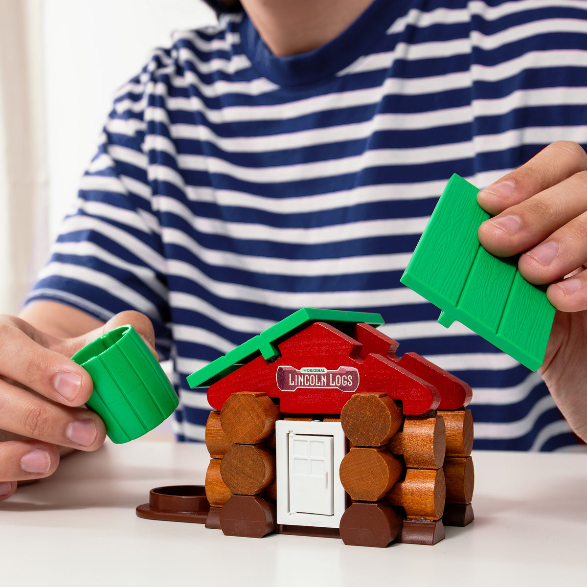 A person wearing a blue and white striped shirt is assembling a small toy log cabin with the brand name "" on the red roof. They are holding green pieces that resemble a cylindrical chimney and part of the roof. The cabin has a white door.