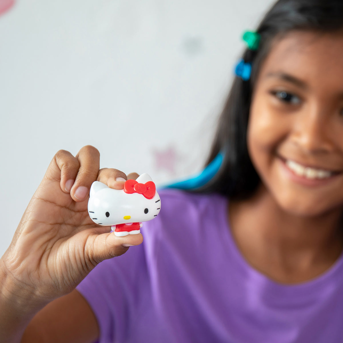 A child with long dark hair, wearing a purple shirt, smiles while holding up a small figurine. The figurine has a white face, red bow, and two black eyes with whiskers. The blurred background decor includes star shapes.