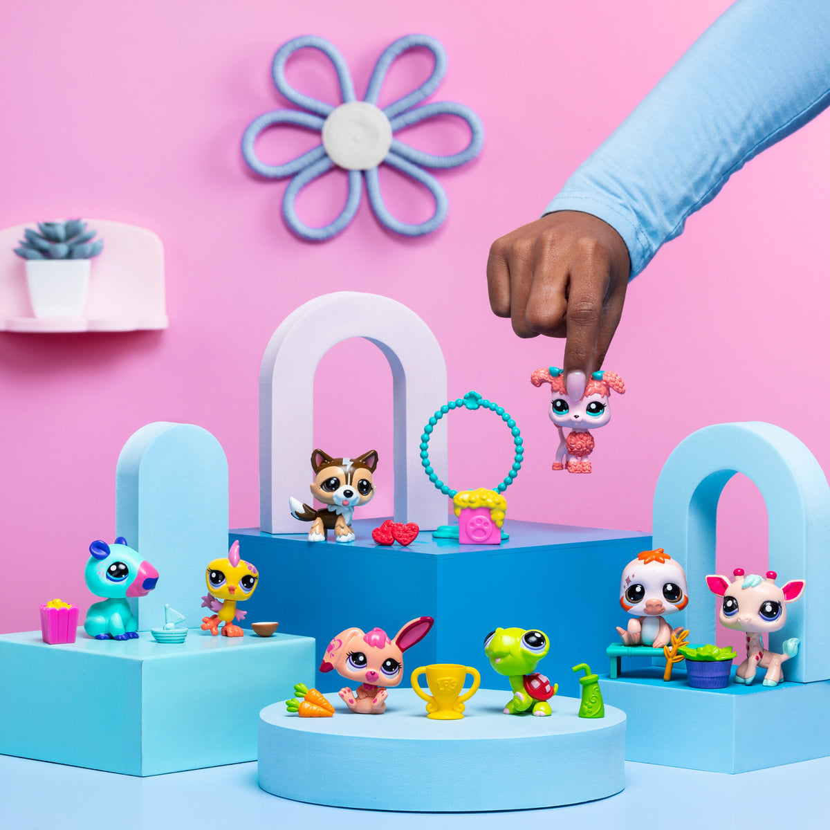A playful scene featuring several '' toy figures arranged on pastel blue and pink geometric platforms against a pink background. A hand is holding one of the pet figures, a pink dog, in mid-air. Other toys include a blue and pink creature with a yellow drink, a yellow bird with a blue cup, a brown dog with red accessories, a green turtle with a trophy, a pink bunny with a carrot, a white and orange puppy, and a white and pink deer-like figure. The background is decorated with a small plant on a shelf and a 