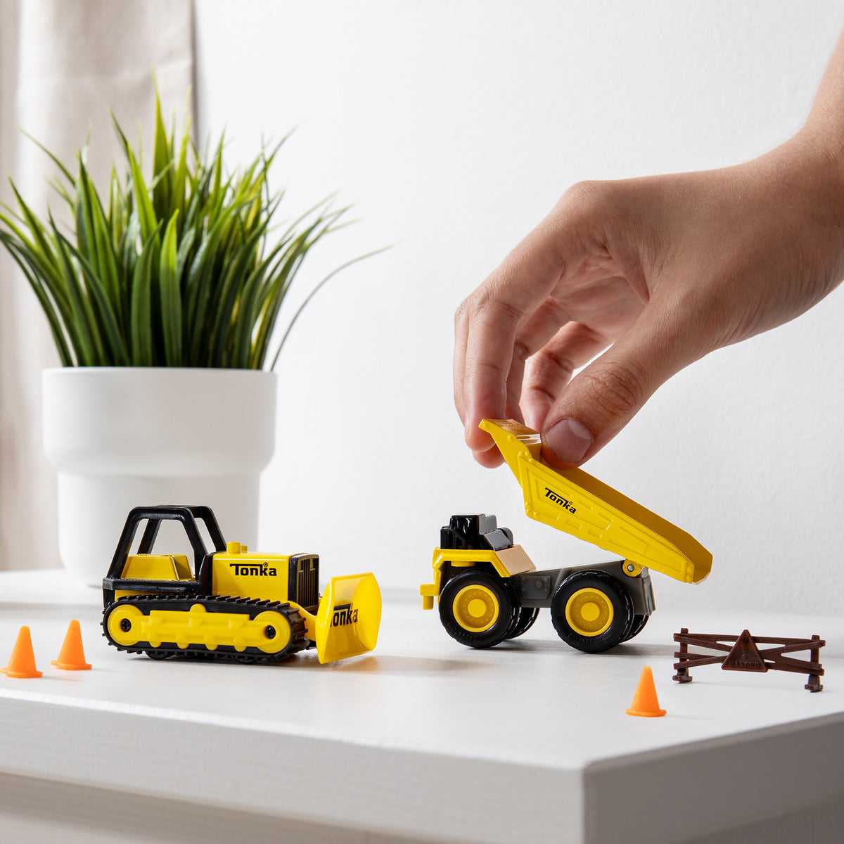 A hand playing with yellow toy construction vehicles on a white surface. One vehicle, with a scoop, faces forward, while the other has its dump bed raised. Small orange cones and a miniature fence are also on the surface. A green potted plant is in the background.